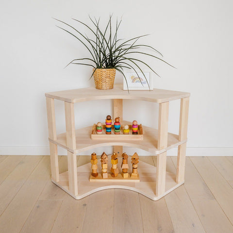 1. Natural wood Montessori corner shelf with two levels, featuring wooden toys and a potted plant on top, placed against a white wall on light wood flooring.