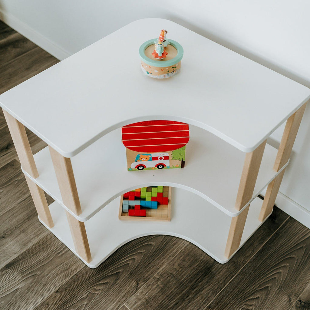 3. Top view of white and natural wood Montessori corner shelf with two levels, displaying a toy house and blocks, on dark wood flooring.