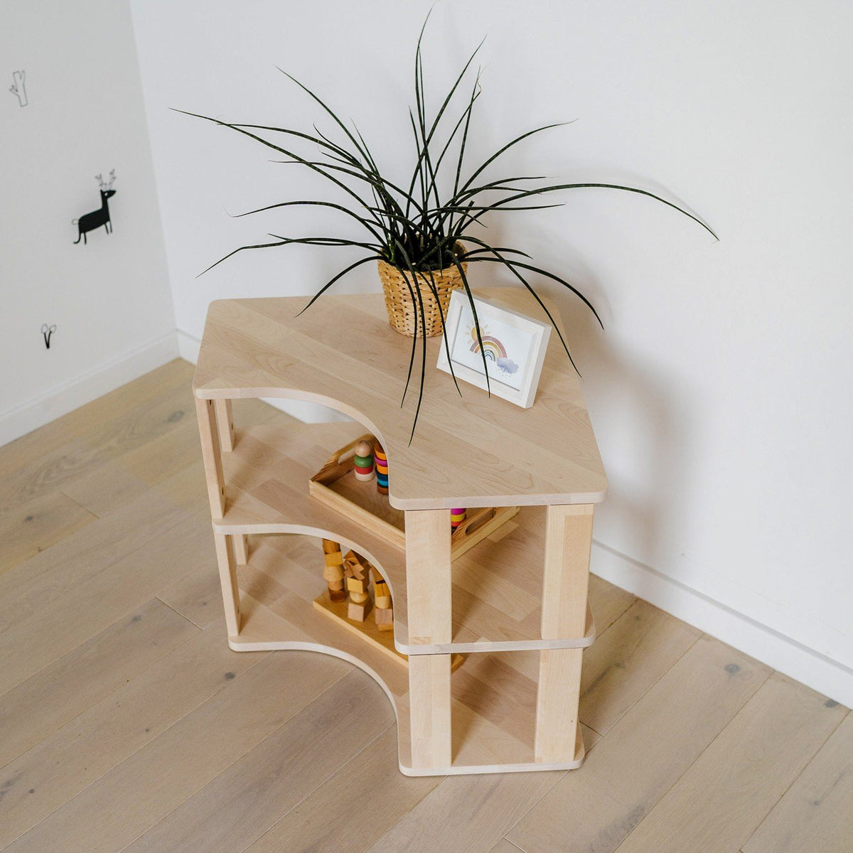 4. Top view of natural wood Montessori corner shelf with two levels, featuring a potted plant and toys, on light wood flooring.