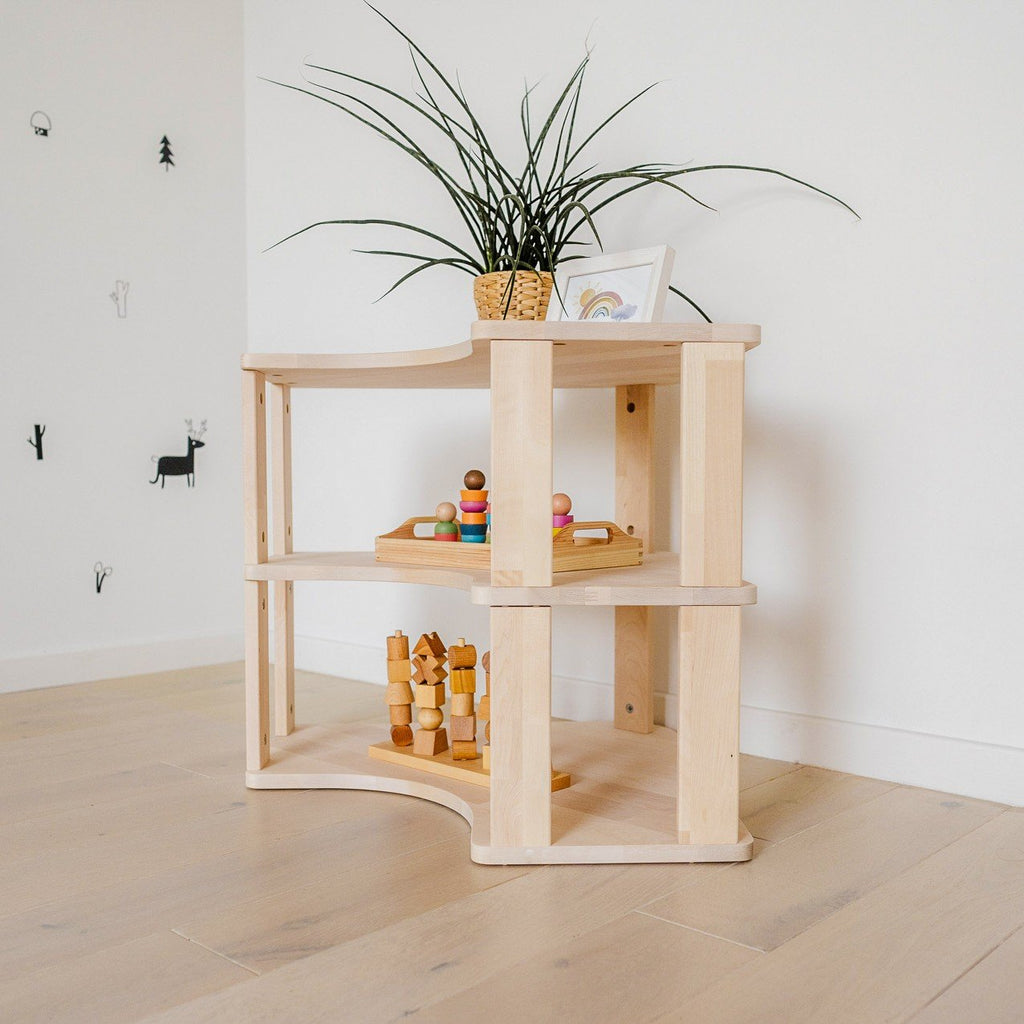 2. Natural wood Montessori corner shelf with two levels, displaying wooden toys and a potted plant, set against a white wall on light wood flooring.
