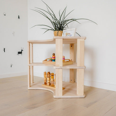 2. Natural wood Montessori corner shelf with two levels, displaying wooden toys and a potted plant, set against a white wall on light wood flooring.