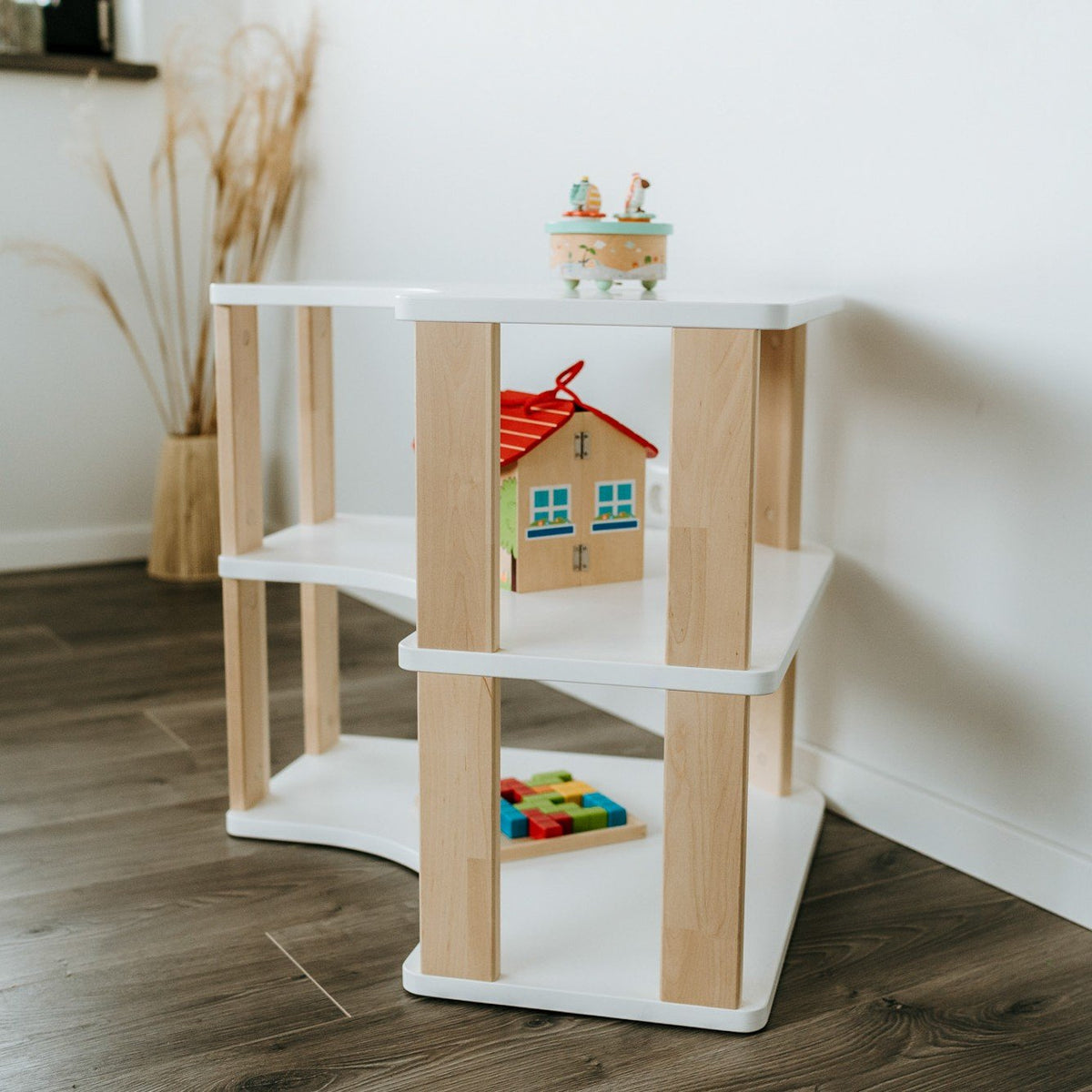3. Side view of white and natural wood Montessori corner shelf with two levels, featuring a toy house and blocks, on dark wood flooring.