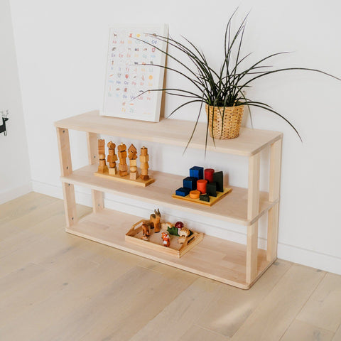 2. Three-tier Montessori shelf with wooden toys and a plant, placed in a minimalist room with light wood flooring