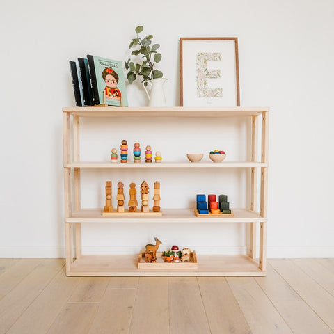 1. Natural wood Montessori shelf with four levels, displaying children's toys and books, set against a white wall with decorative items on top