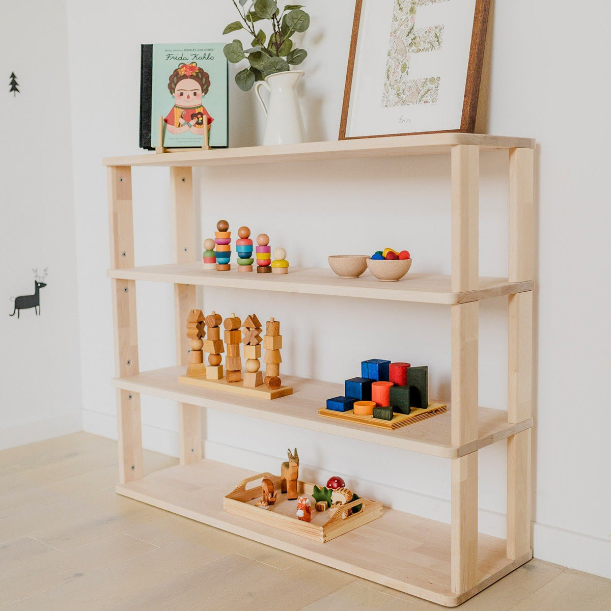 3. Side view of natural wood Montessori shelf with four levels, displaying colorful toys and books, against a white wall