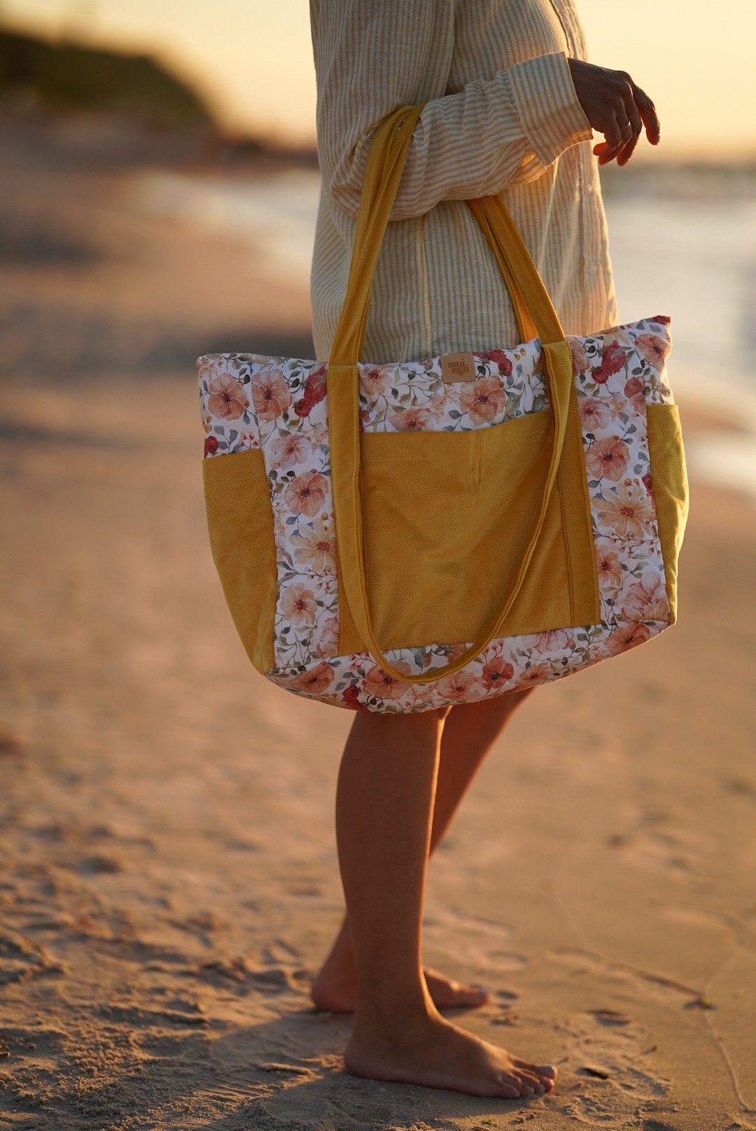 1. Woman holding yellow and floral weekender bag on sandy beach at sunset