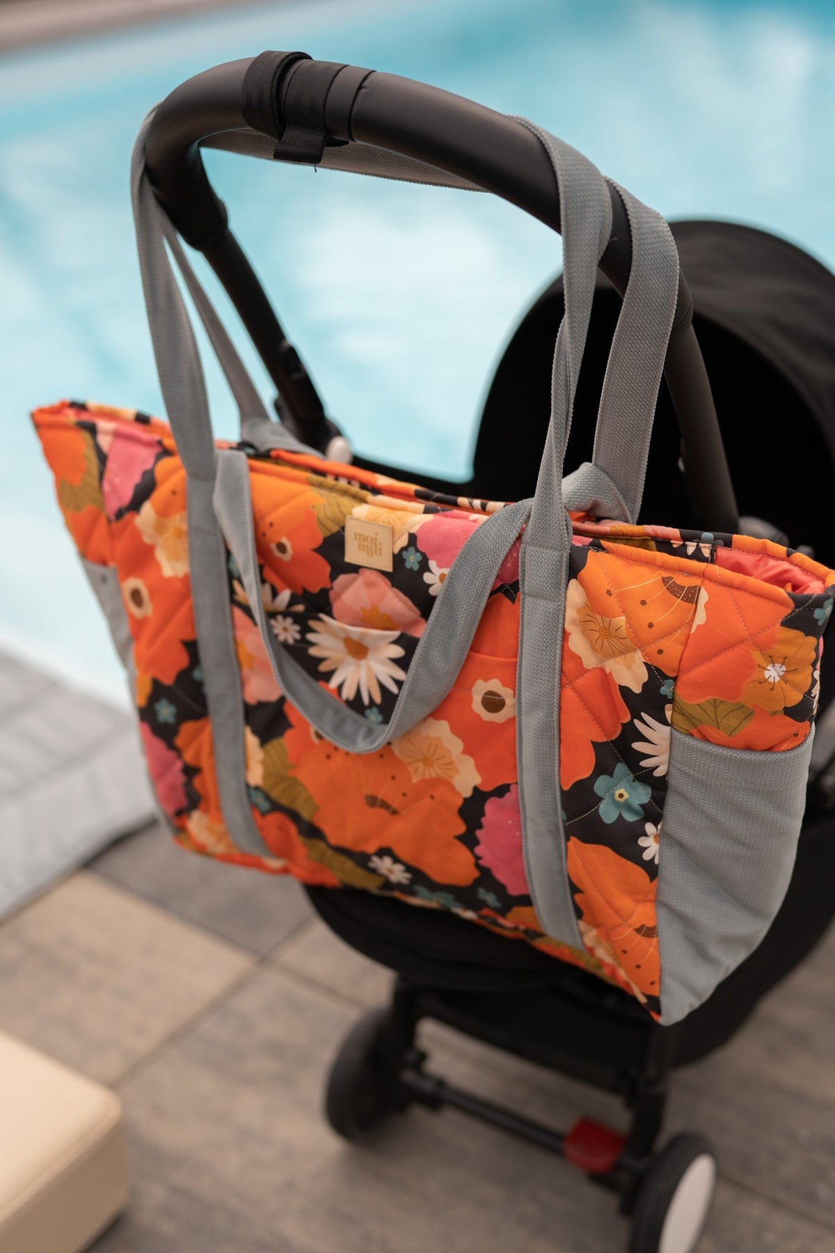 1. Floral weekender bag with blue straps hanging on a stroller by a poolside, showcasing its versatility for parents.