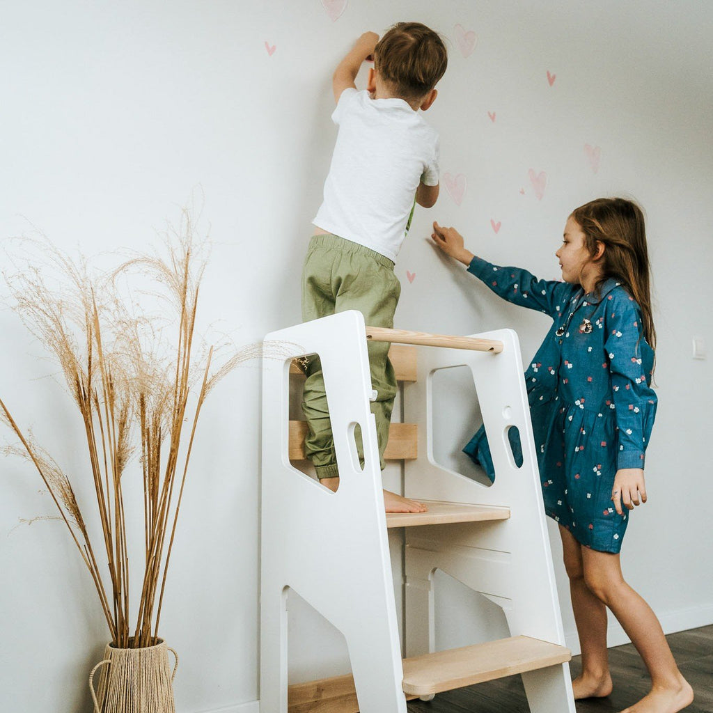 1. Two children using white Montessori Tower Safety+ to reach wall decorations, highlighting its safety and collaborative use