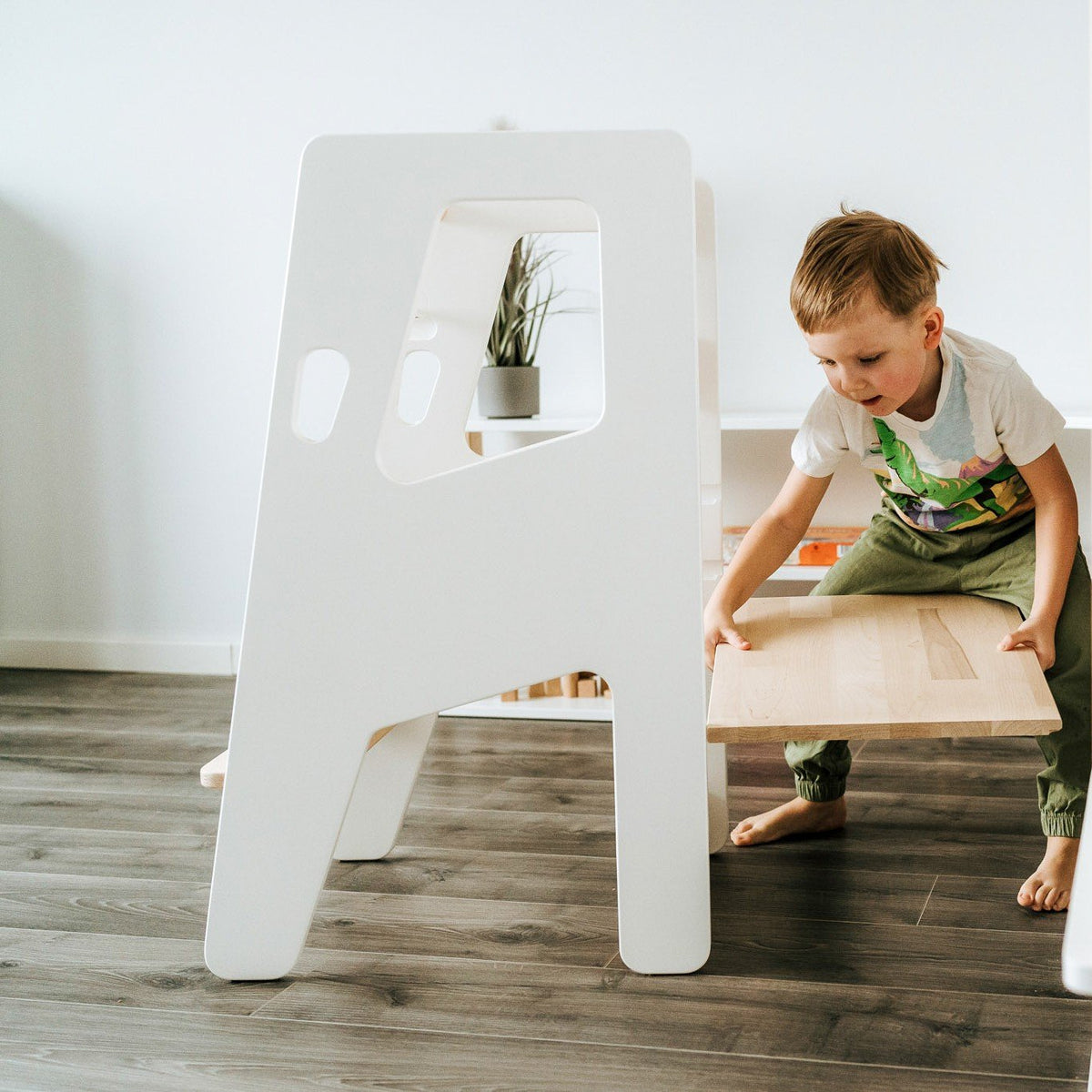 1. Child adjusting the platform of a white Montessori Tower Safety+ in a bright room, illustrating its adjustable height feature