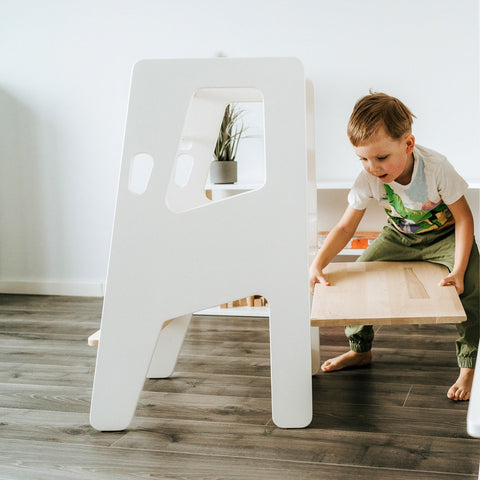 1. Child adjusting the platform of a white Montessori Tower Safety+ in a bright room, illustrating its adjustable height feature