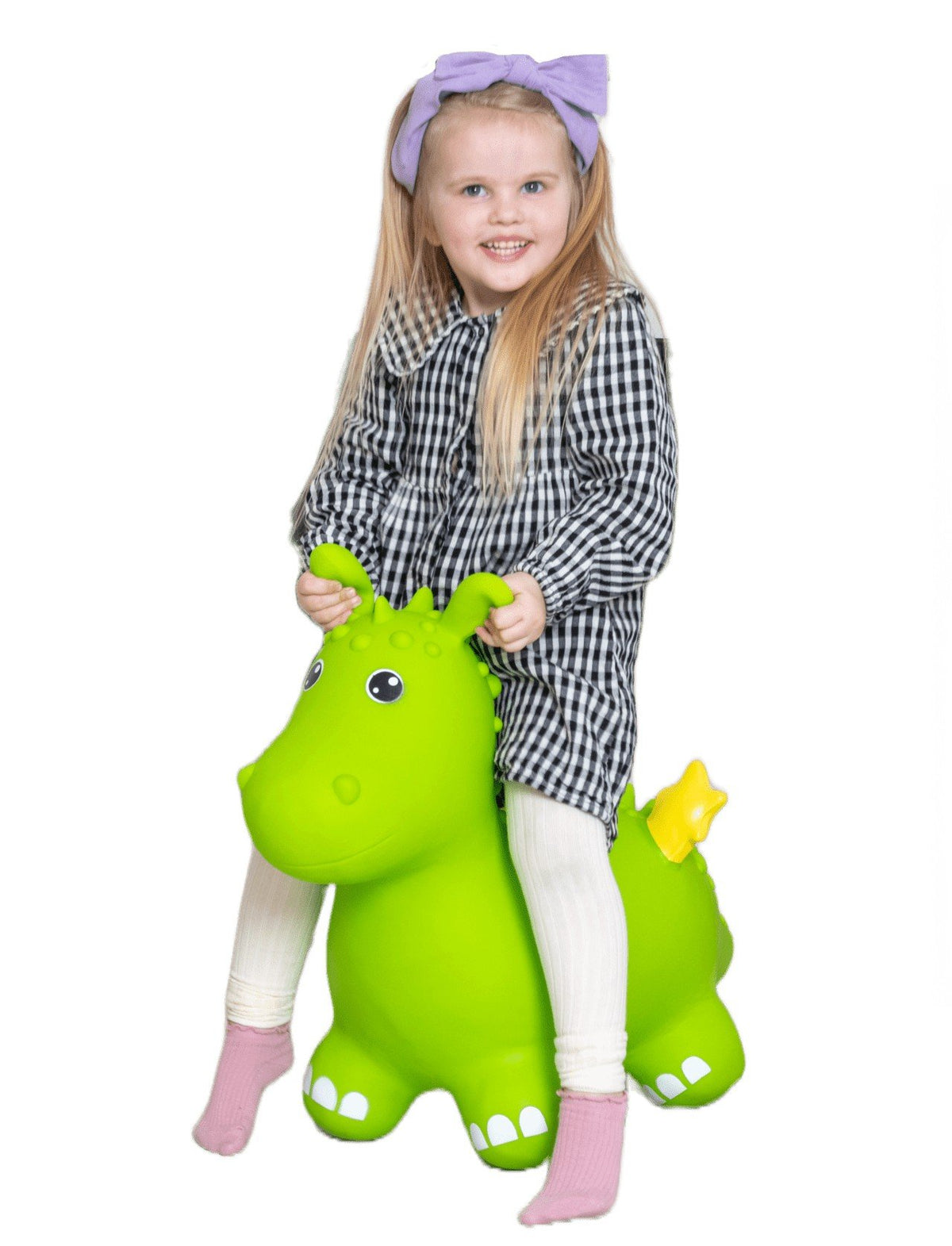 1. Young girl with blonde hair and purple bow riding a green dragon-shaped hopper toy indoors, wearing a black and white checkered dress and pink socks