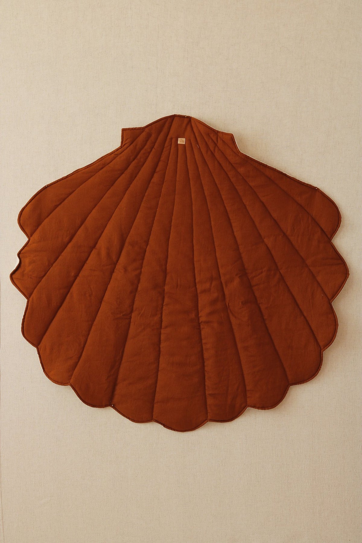 2. Caramel shell-shaped playmat hanging on a beige wall, highlighting its quilted design and scalloped edges