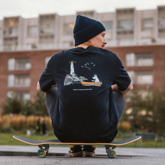 1. Unisex Moomin by NordicBuddies T-shirt worn by male model sitting on skateboard, showing back design with lighthouse and Moominpappa illustration