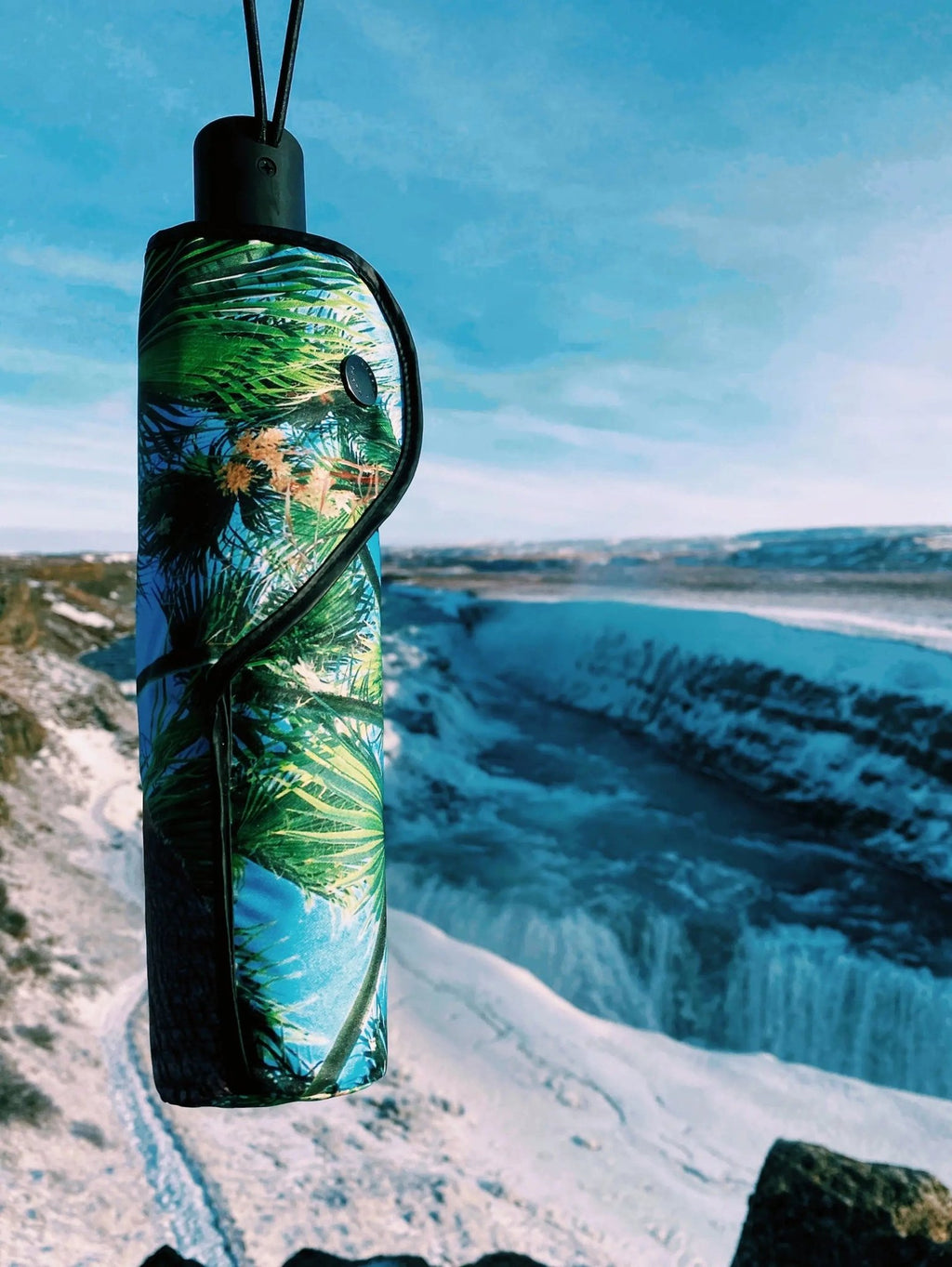 4. Closed Paradise Umbrella with tropical print, hanging against a scenic waterfall background