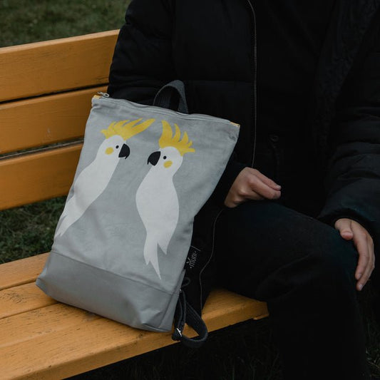 2. Muni Cockatoo backpack in grey velvet with cockatoo design, placed on a bench next to a person in a black jacket