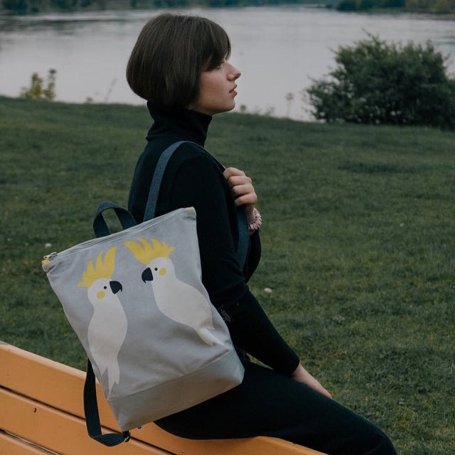 1. Woman sitting on a bench with Muni Cockatoo backpack in grey velvet, featuring cockatoo design, in a park setting