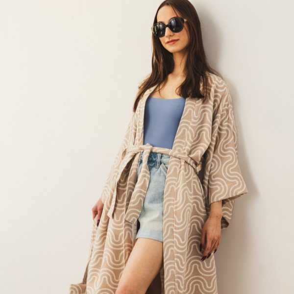 1. Woman wearing Patulove sand bamboo kimono with blue top and denim shorts, leaning against a white wall