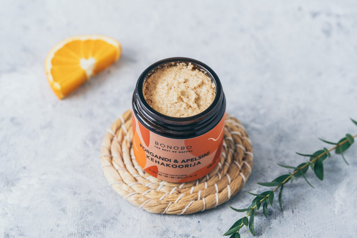 3. Open jar of BONOBO Orange & Carrot body scrub on a woven mat with orange slice and greenery