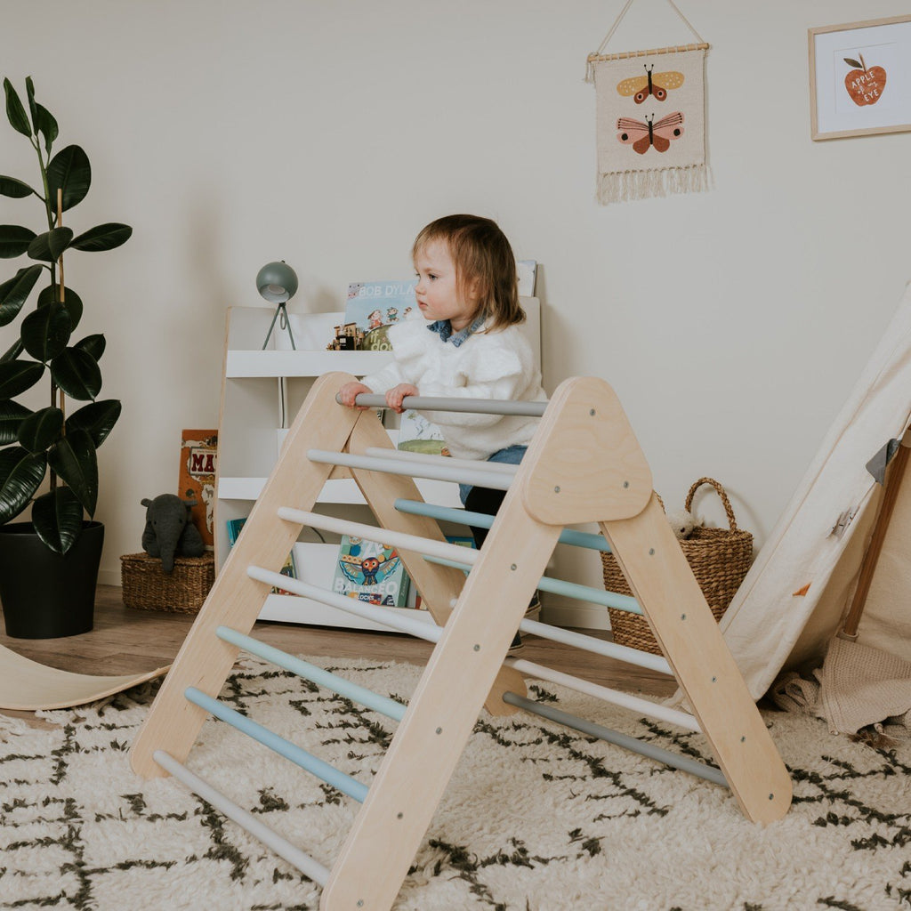1. Child standing on pastel Pikler Triangle in cozy playroom with books and plant