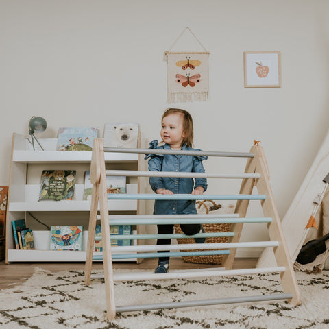 3. Child in denim outfit climbing pastel Pikler Triangle in playroom with books