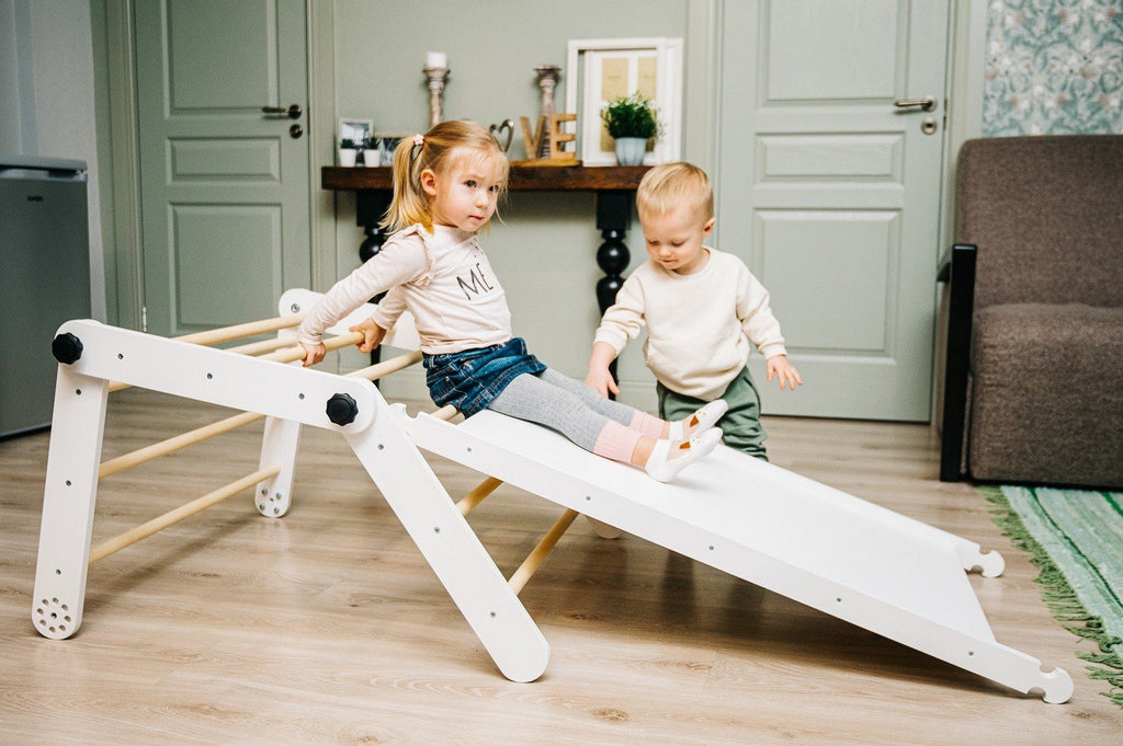 4. Girl sliding down a white Pikler triangle with a boy standing nearby in a living room