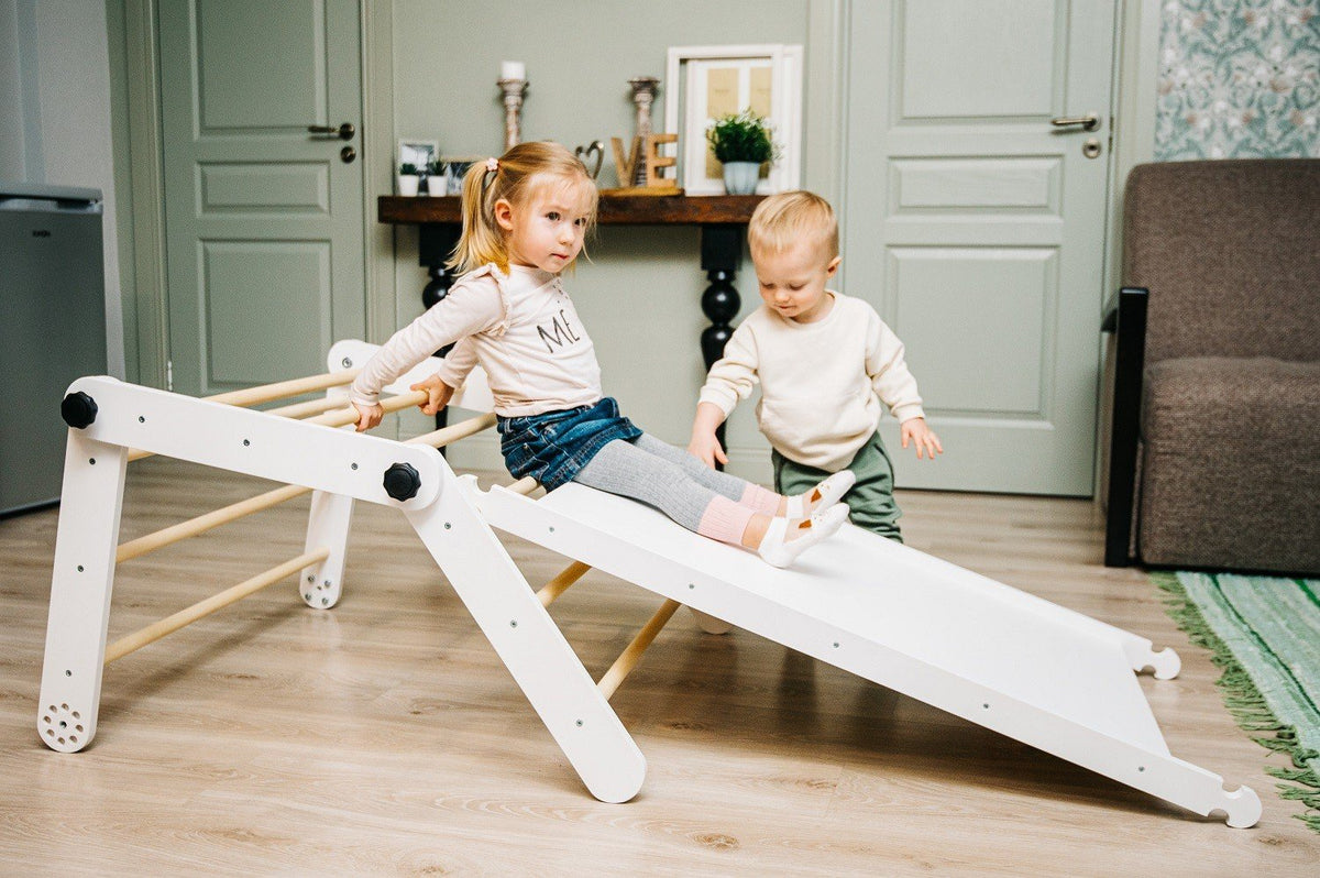 1. Two children playing on a white Family-scl Pikler Triangle with ramp in a living room setting, featuring birch plywood construction and climbing rocks, suitable for ages 2 to 7.