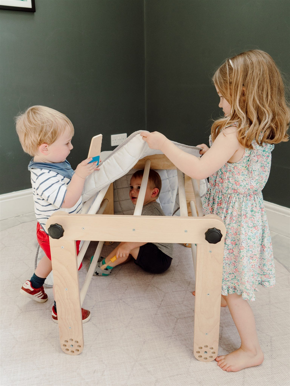 1. Two children playing with a Pikler triangle covered with a blanket, one child inside and one outside, in a playroom setting