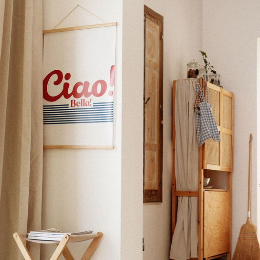 1. 'Ciao Bella' poster with red typography and blue stripes hanging in a cozy room with wooden furniture and neutral curtains