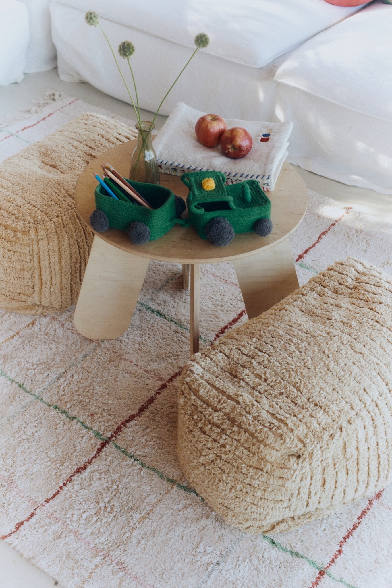 4. Flat lay of Lorena Canals Pouf Haystacks in light honey, arranged around a small wooden table with toys and decor, on a patterned rug