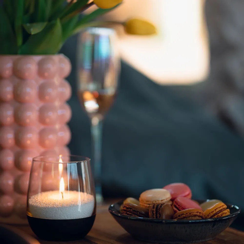 1. Lit candle in glass holder on table with pink vase, champagne glass, and macarons in cozy setting