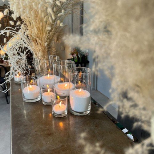 1. Multiple glass candles with white sand and flames on a wooden surface surrounded by dried floral decor