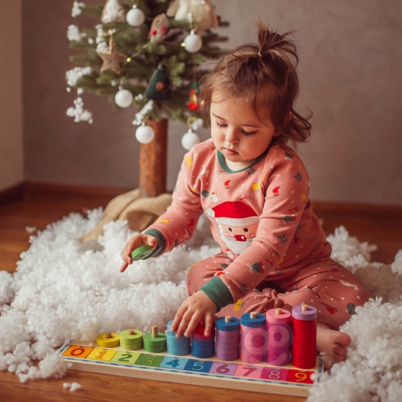 6. Child in festive pajamas arranging colorful rings on a wooden counting puzzle near a Christmas tree