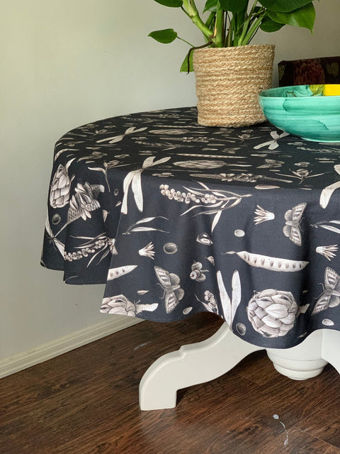 4. Black Seeds Carbon print tablecloth on a round table, featuring artichoke and pea pod designs, with a green plant and turquoise bowl in a dining room