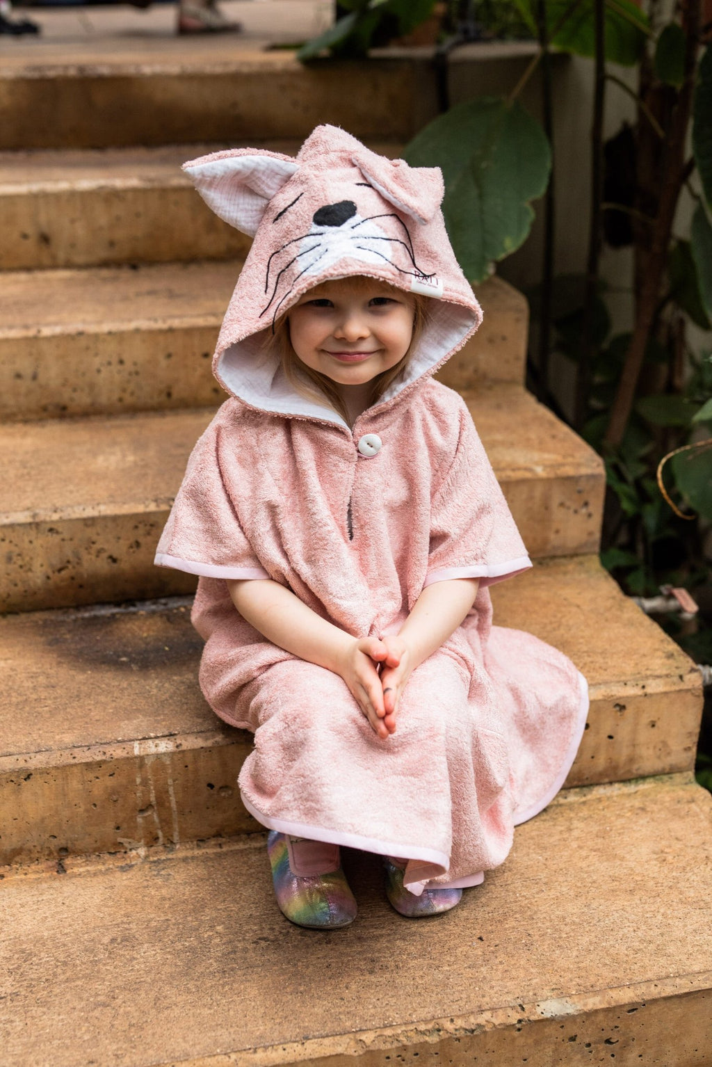 2. Smiling child sitting on steps wearing RÄTT Short-Sleeve Poncho CAT with hood, showcasing cozy fit