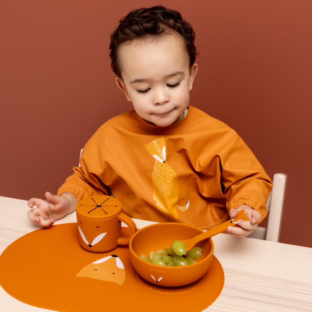 1. Child wearing orange Trixie Baby waterproof long sleeve bib with Mr. Fox design, sitting at table with matching fox-themed dinnerware