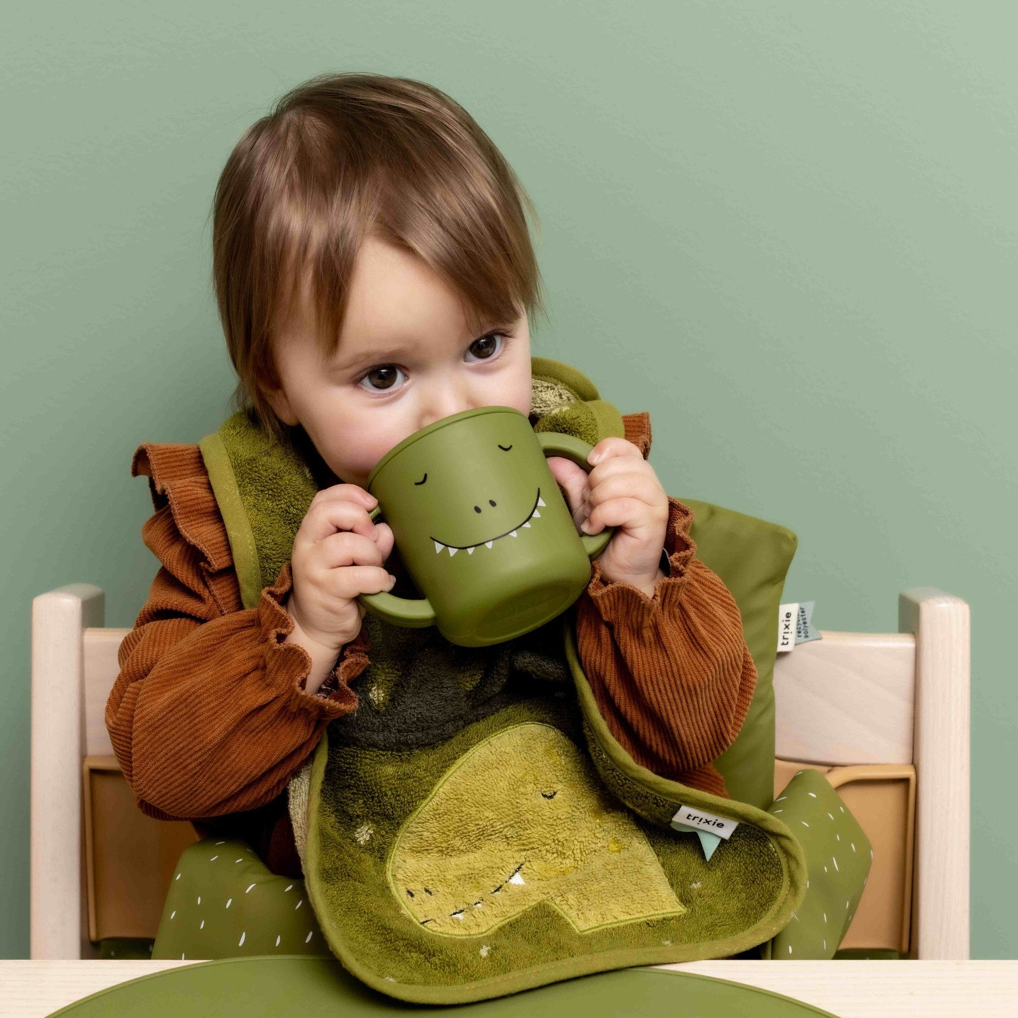 1. Toddler drinking from green dinosaur-themed silicone sippy cup at table