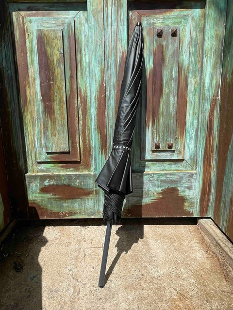 2. Closed black HAPPYSWEEDS parasol leaning against rustic wooden door highlighting brand logo