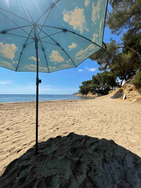 1. Beach scene with open HAPPYSWEEDS parasol featuring sky and cloud design providing shade on sandy shore with sea and trees in background