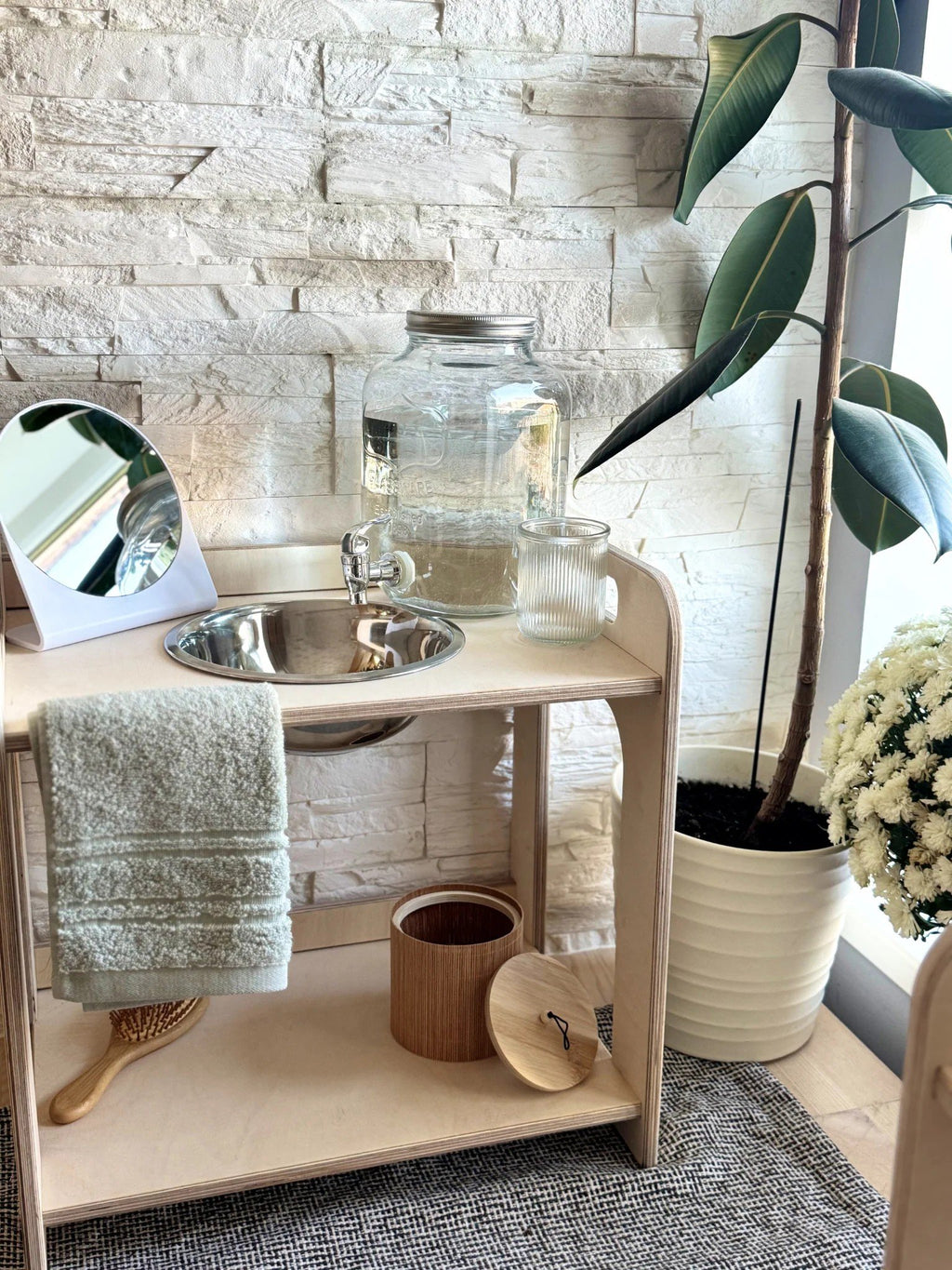 1. Montessori wooden wash station with enamel bowl, towel, and glass jar in a natural setting with stone wall and plant