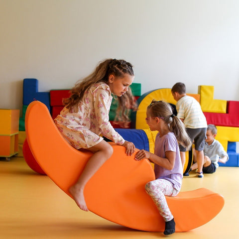 1. Two girls playing on orange soft play double rocker in playroom with other children in background, wearing casual outfits