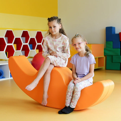 1. Two girls sitting on orange soft play double rocker in playroom, one wearing floral dress and the other in purple top