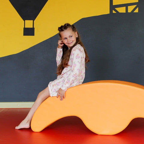 1. Girl sitting on orange soft play double rocker in playroom with colorful wall art, wearing floral dress