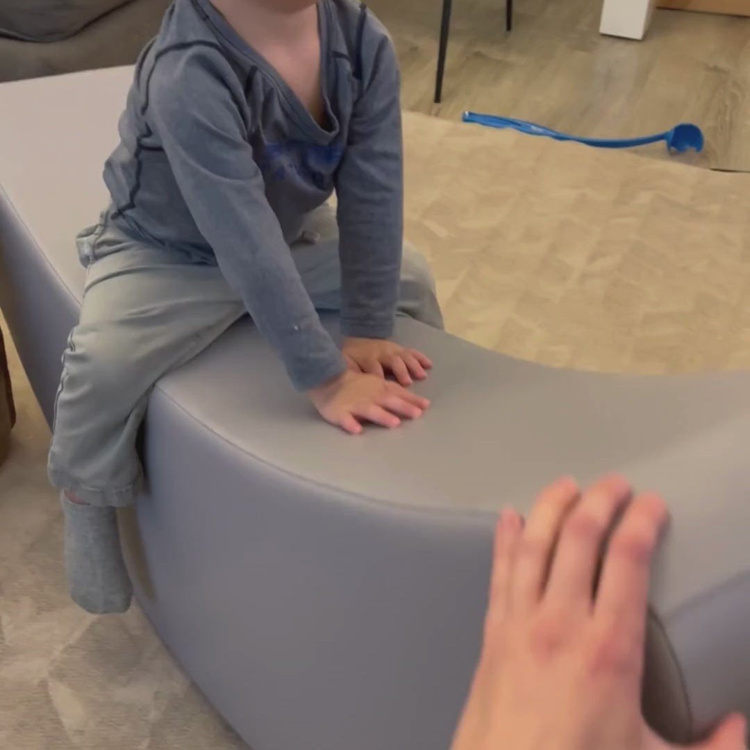 9. Child sitting on grey banana-shaped rocking toy indoors with adult hand visible