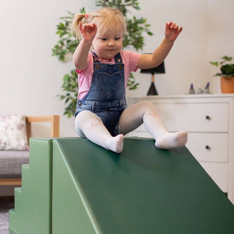 4. Child in denim overalls sliding down green soft play set in a home environment