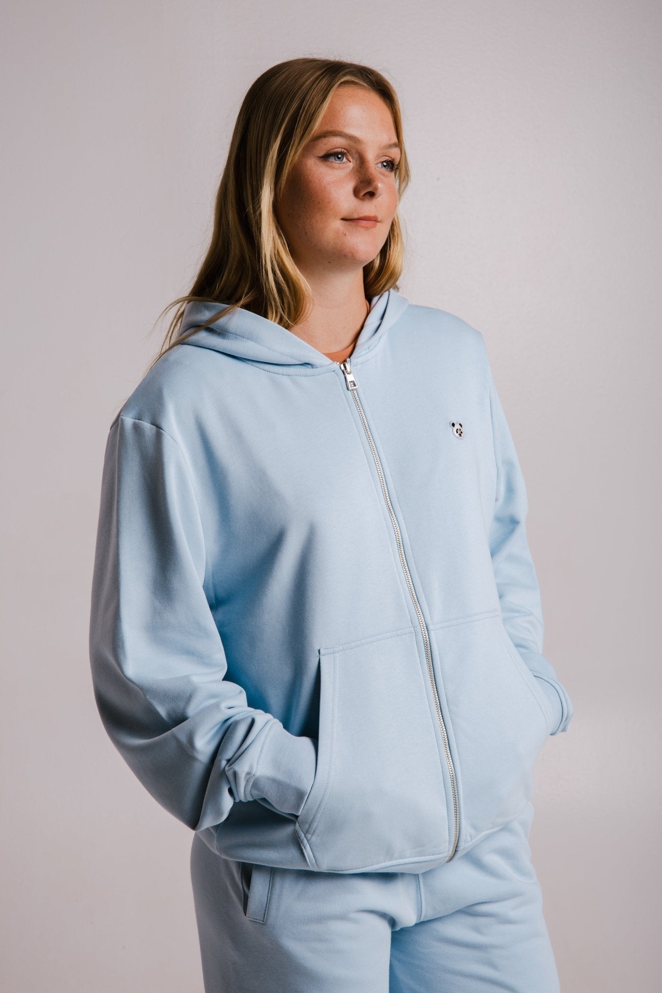 2. Woman in Sossu light blue Panda Soft Fleece Full Zip Hoodie with hands in pockets, studio shot