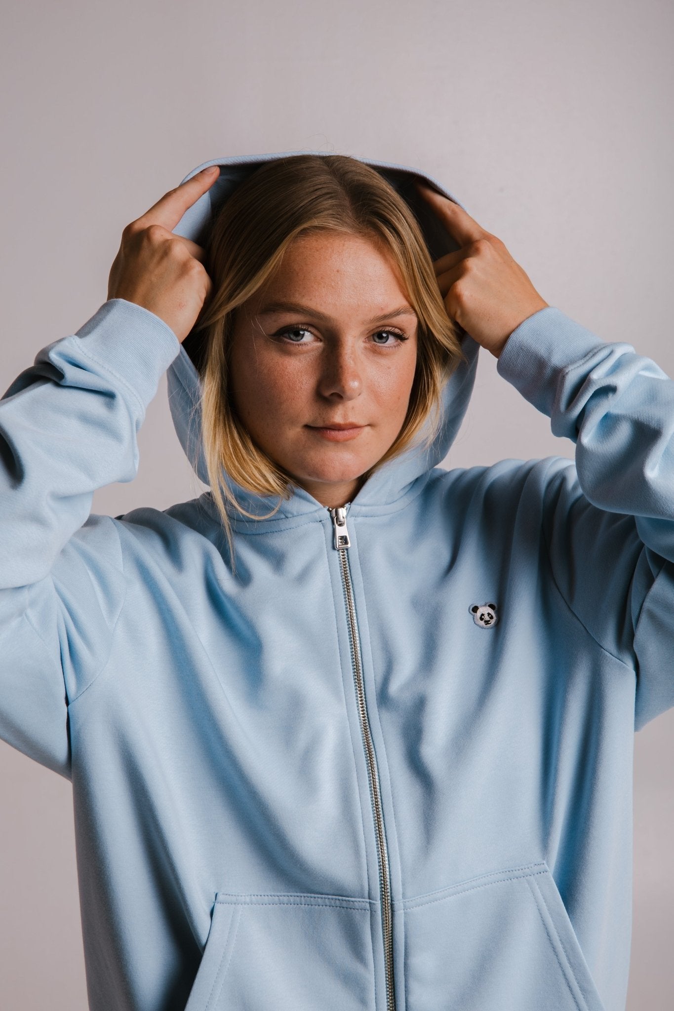 1. Woman wearing Sossu Panda Soft Fleece Full Zip Hoodie in light blue, adjusting hood in studio setting