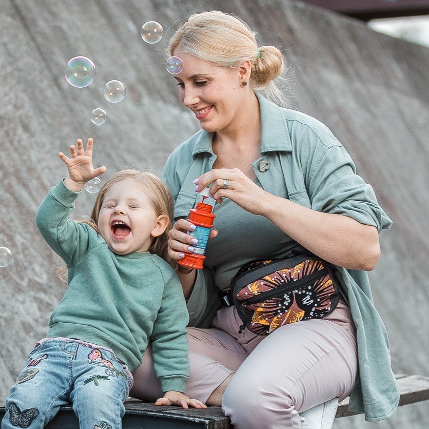 1. Woman wearing Drinbags waist bag with kaleidoscope pattern, playing with child blowing bubbles outdoors