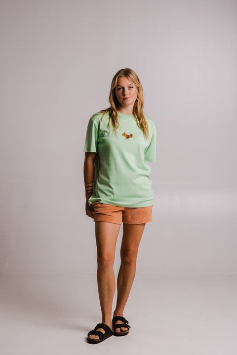 2. Full view of woman wearing Sossu Squirrel Heavy T-shirt in light green, paired with orange shorts in studio