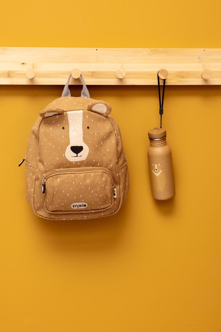 9. Trixie Baby Mr. Bear backpack and matching stainless steel bottle hanging on wooden hooks, against yellow background
