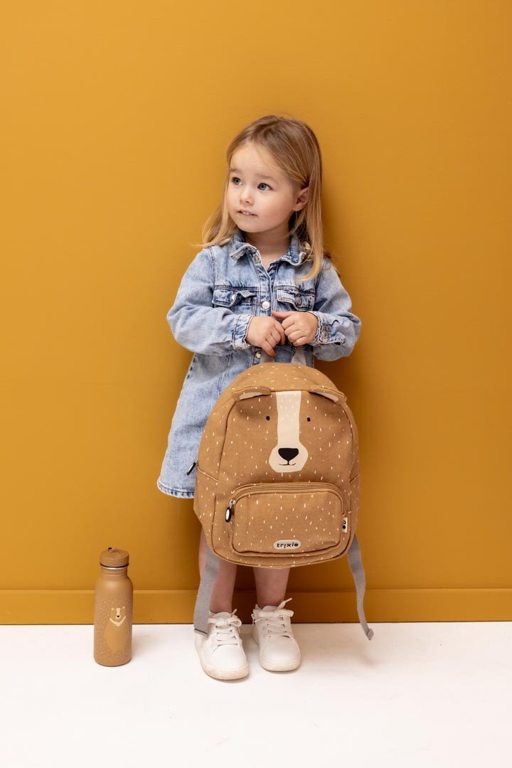 8. Young girl in denim dress with Trixie Baby Mr. Bear backpack and matching stainless steel bottle, against yellow background